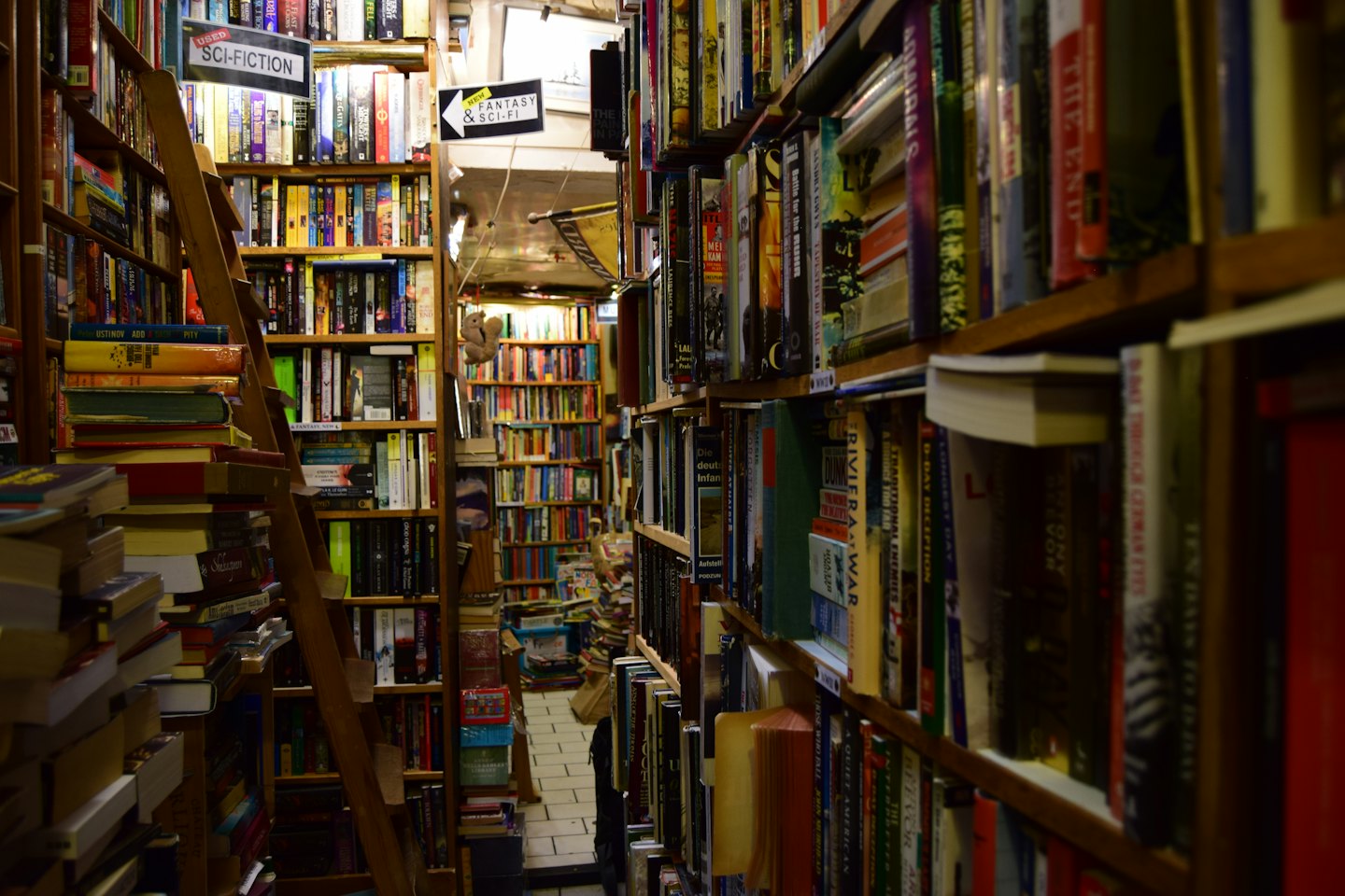 Stretto corridoio in una libreria dell'usato con scaffali traboccanti e una scala a rotelle.